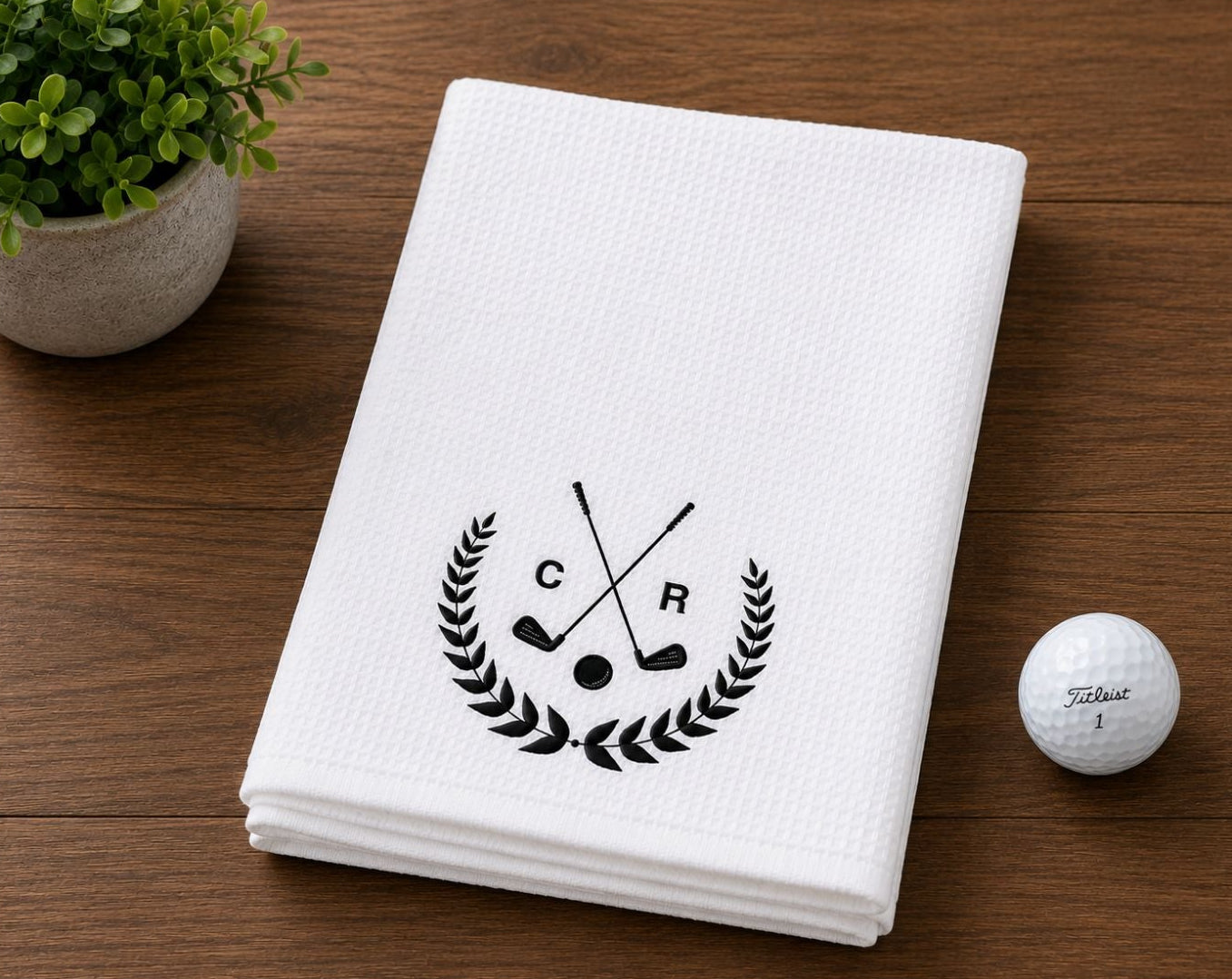 White towel with golf club logo and custom initials on a wooden surface with a golf ball and plant.